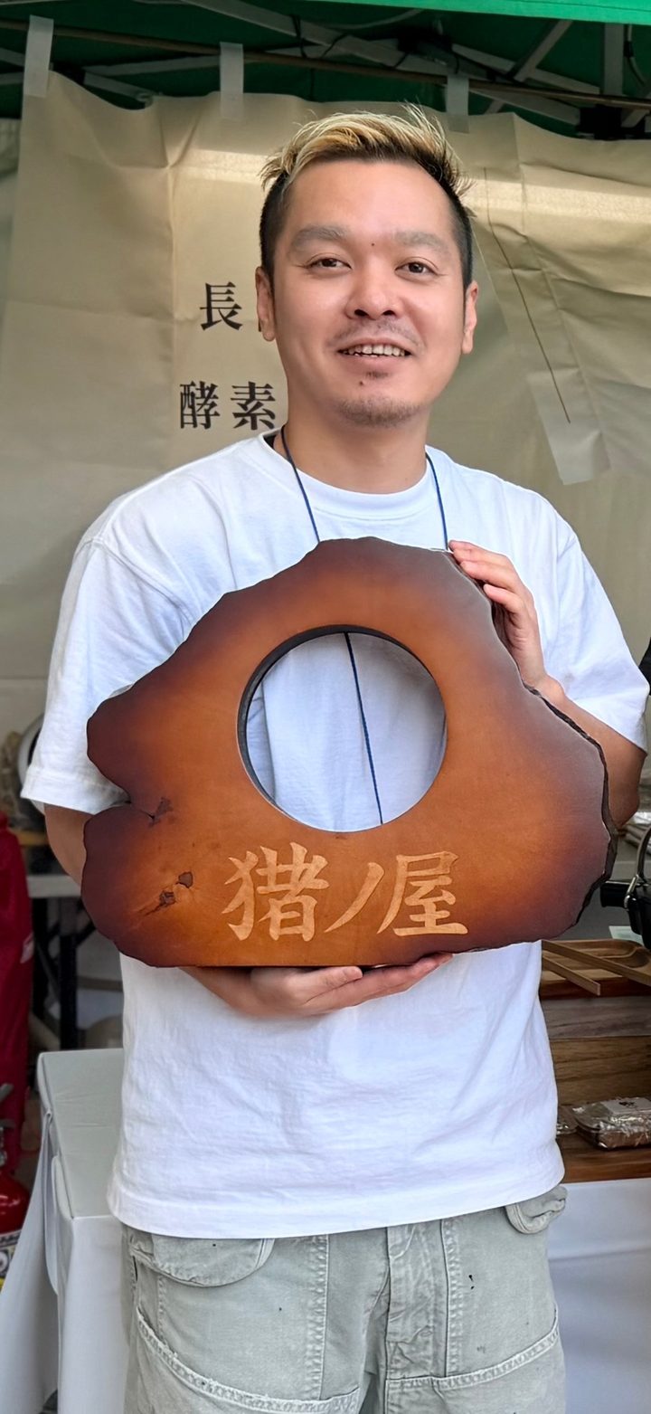 合同会社猪ノ屋 代表 服部俊喜。長岡式酵素玄米のブースで、木製の看板を手に微笑んでいる。